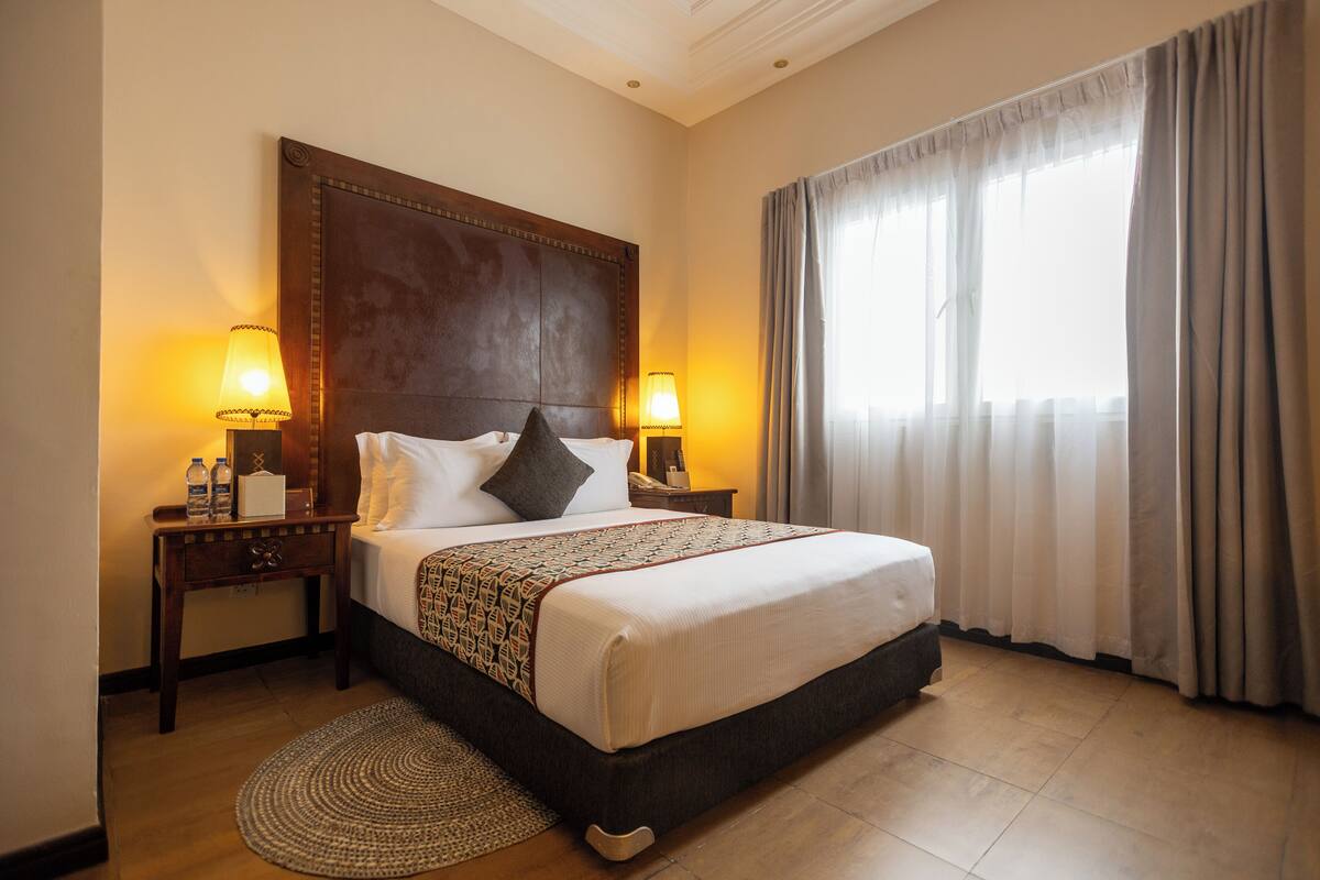 A hotel room with a large bed, two bedside lamps, a wooden headboard, a round rug, and a window with curtains.