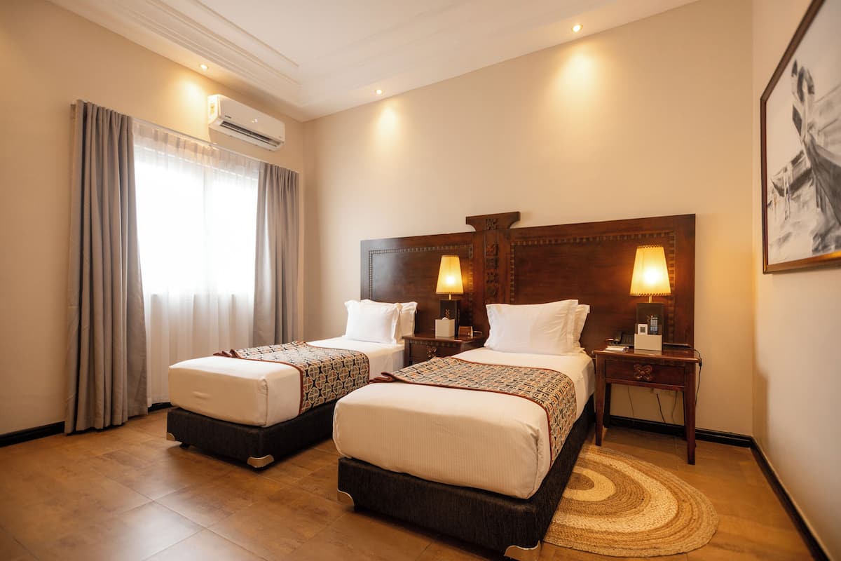 A hotel room with two beds, a wooden headboard, bedside lamps, a bedside table, and a large window with curtains.