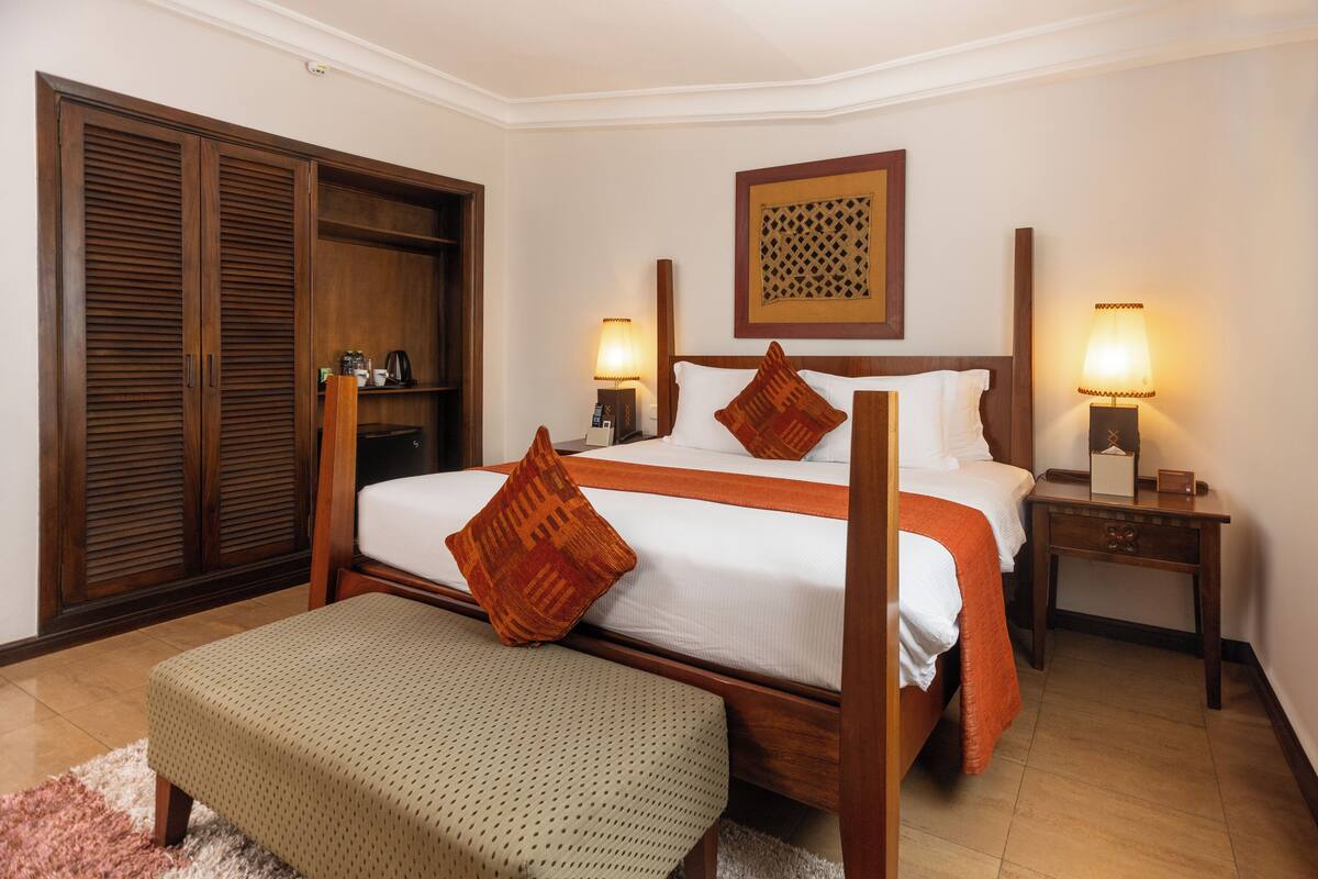 A hotel room with a large bed, a wooden wardrobe, a bedside table with a lamp, and a bench.