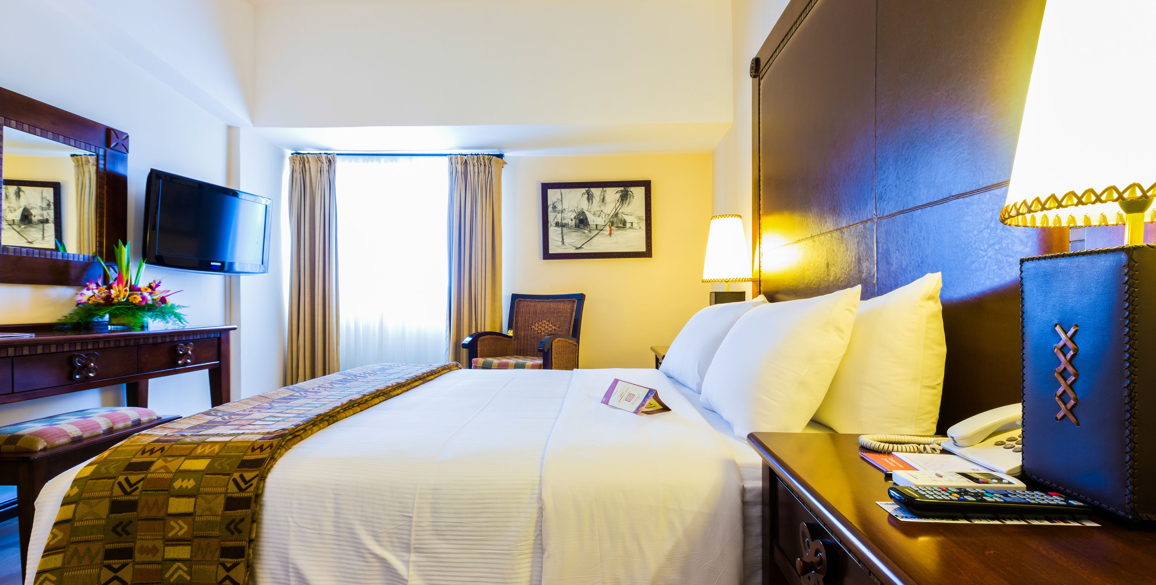 A hotel room with a large bed, a desk with a lamp, a chair, a TV, and a window with curtains.