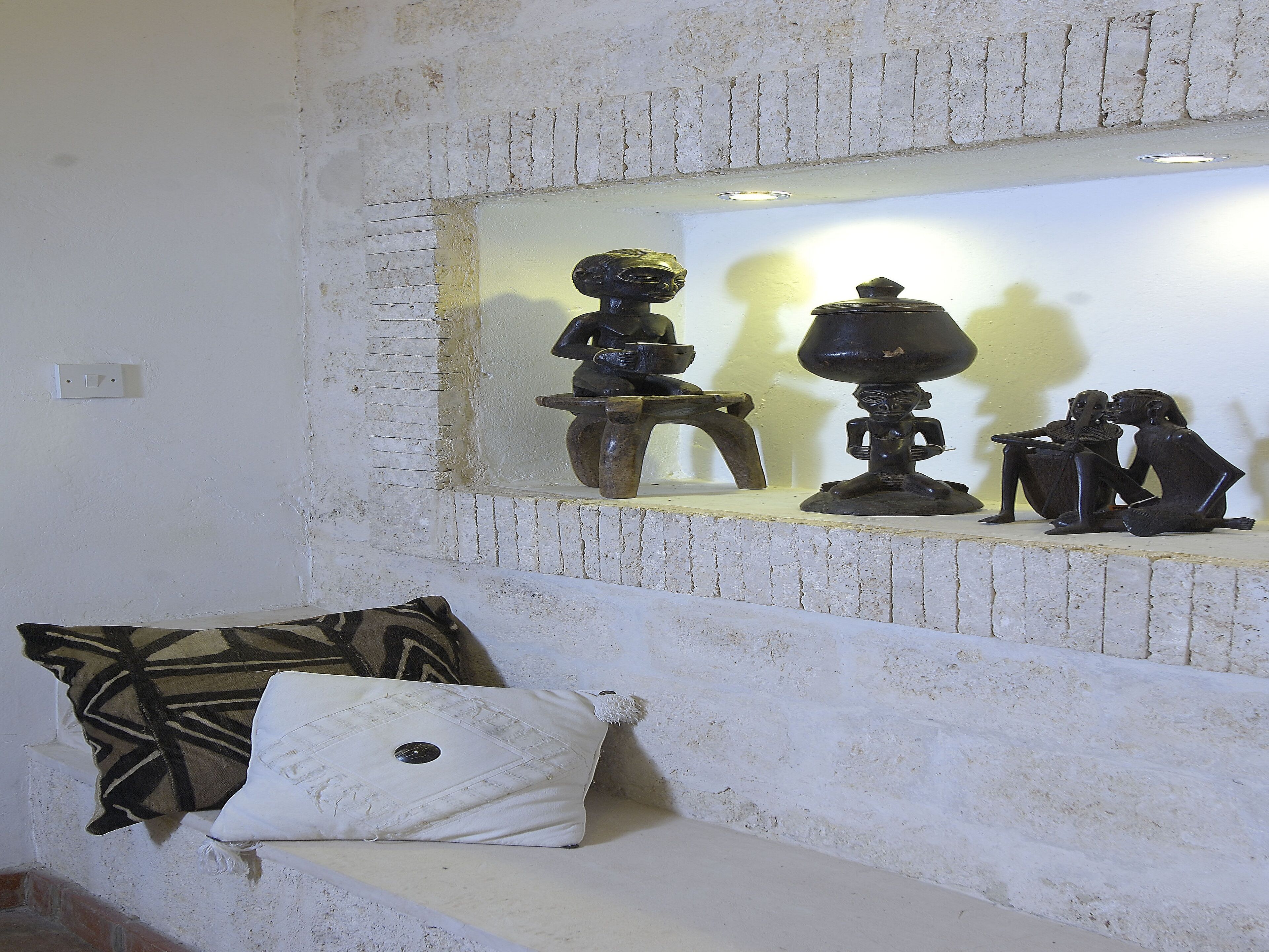A room with a white brick wall, a shelf with three black sculptures, and a white cushion with a geometric pattern on a bench.
