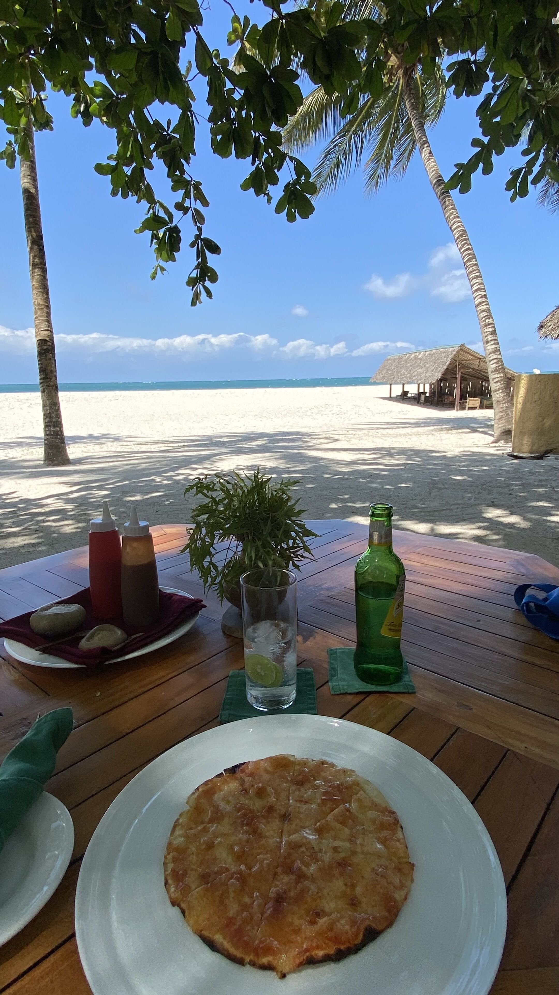 Una mesa de madera con un plato de comida, salsas, un vaso de agua y una botella, con vistas a una playa con palmeras y cielo despejado.