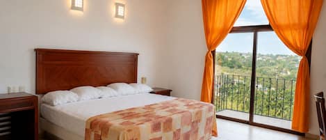 Un dormitorio con cama, mesitas de noche, ventana con cortinas y vistas al verdor.