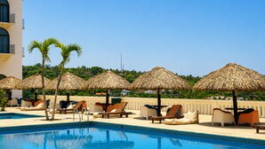 Zona de piscina con sillones de descanso y sombrillas de paja, rodeada por un edificio con balcones.