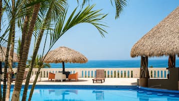A poolside area with a view of the ocean, palm trees, and thatched umbrellas.