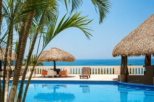 Zona de piscina con vista al océano, palmeras y sombrillas de paja.