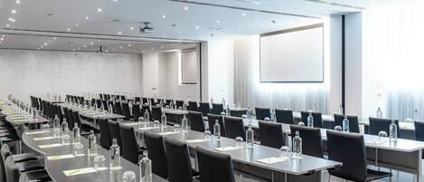 A conference room with rows of tables, chairs, and water bottles, equipped with a projector screen and a large window.