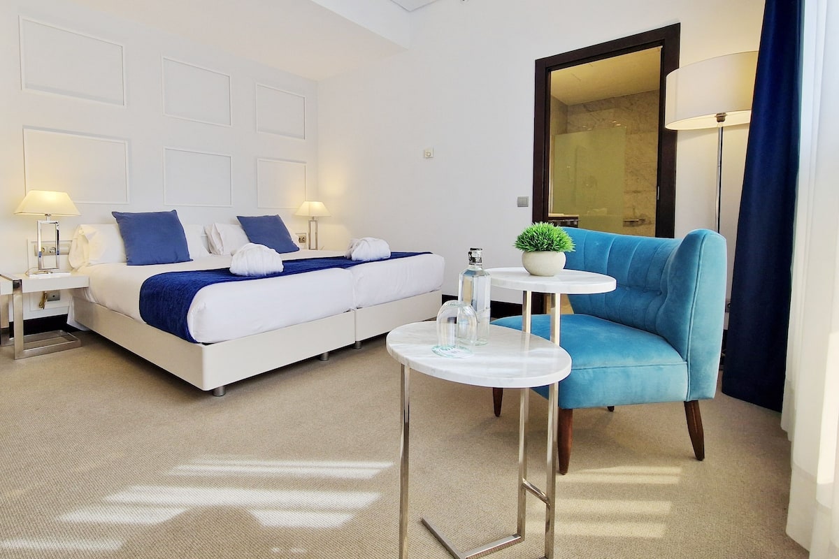 A modern hotel room with a bed, a blue armchair, a small table with a plant, and a mirror.