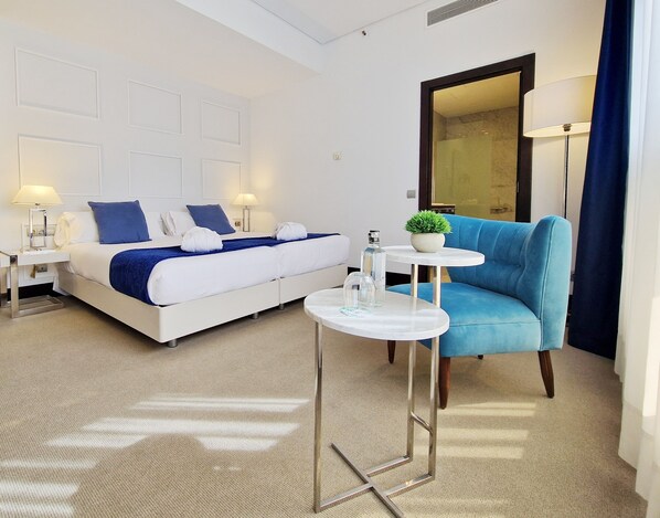 Una habitación de hotel moderna con una cama, un sillón azul, una mesita con una planta y un espejo.