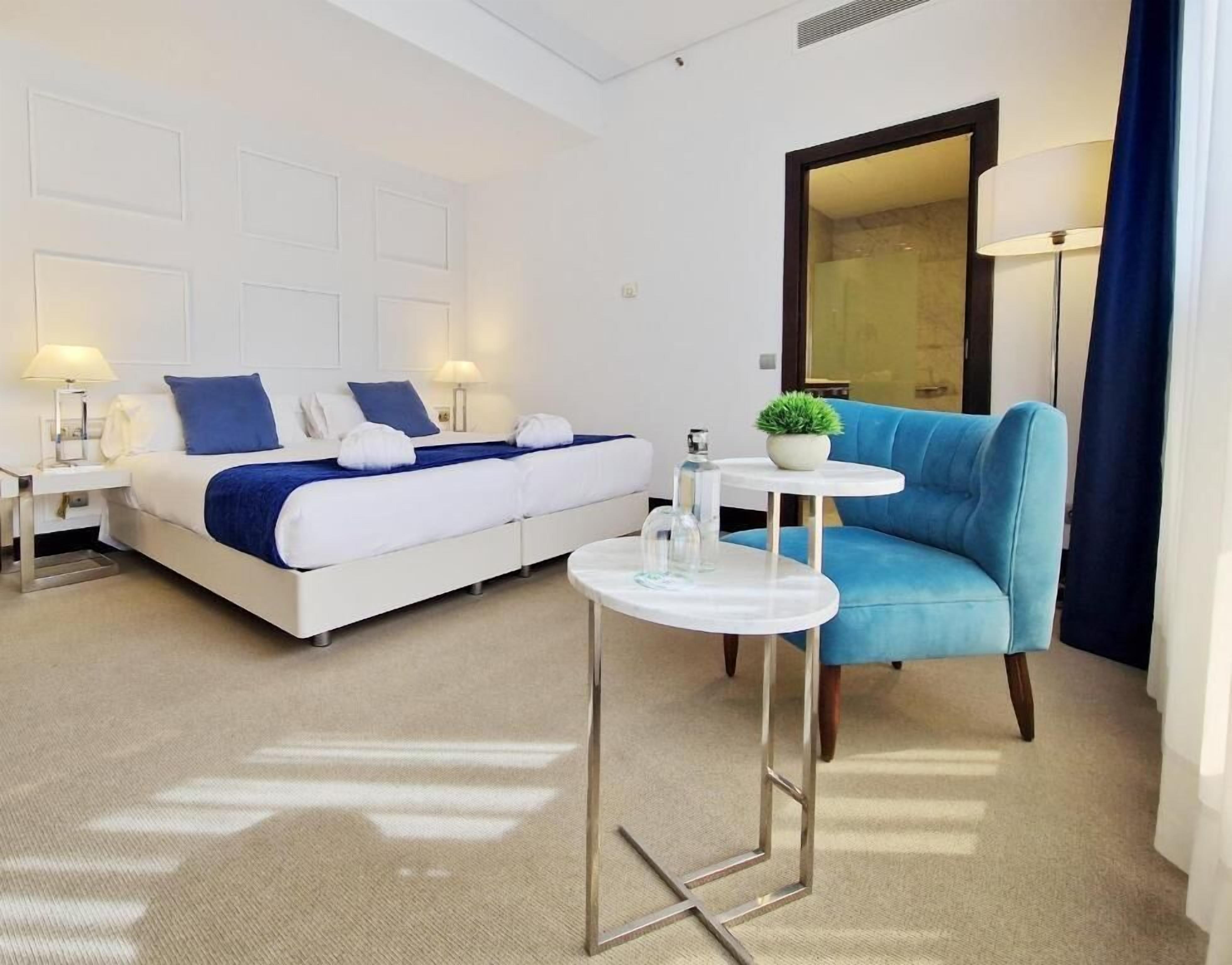 Una habitación de hotel con una cama, un sillón azul, una mesita con una planta y un espejo.