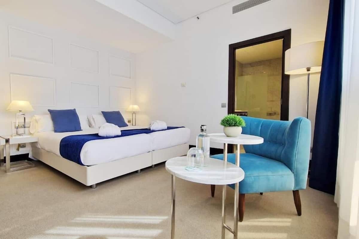 Una habitación de hotel con una cama, un sillón azul, una mesita con una planta y un espejo.