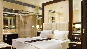 A hotel room with a large bed, a bathroom with a shower, a vanity with a mirror, and a nightstand with a lamp. - Hotel Emperor Palms at Karol Bagh (New Delhi)