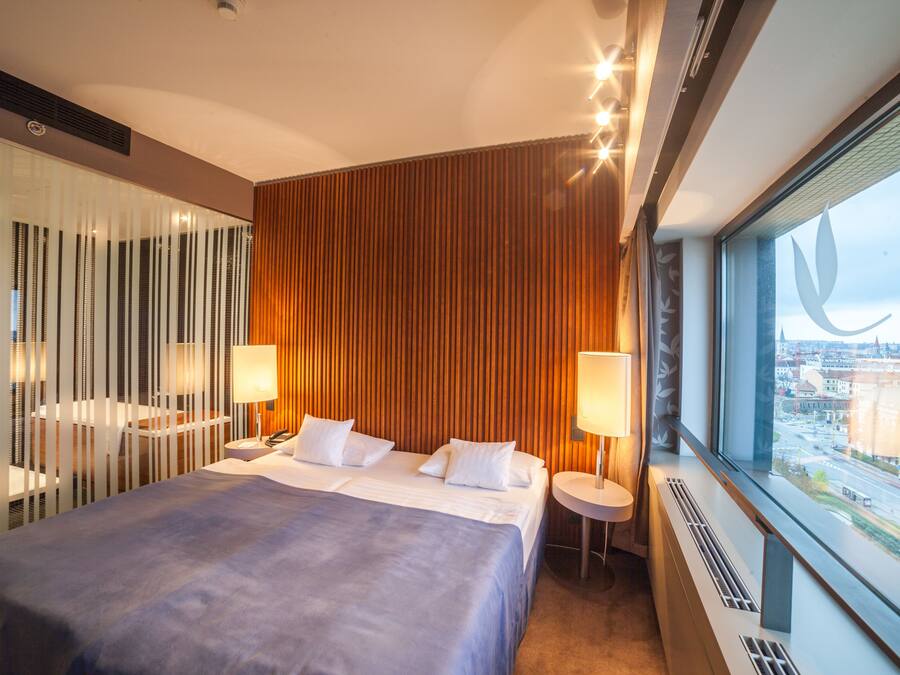 Una moderna camera d'hotel con un letto grande, due comodini con lampade, vista sulla città attraverso un'ampia finestra e una parete rivestita in legno.