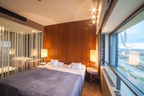 A modern hotel room with a large bed, two bedside tables with lamps, a view of the city through a large window, and a wooden panel wall. - Hotel Yasmin (Kosice)