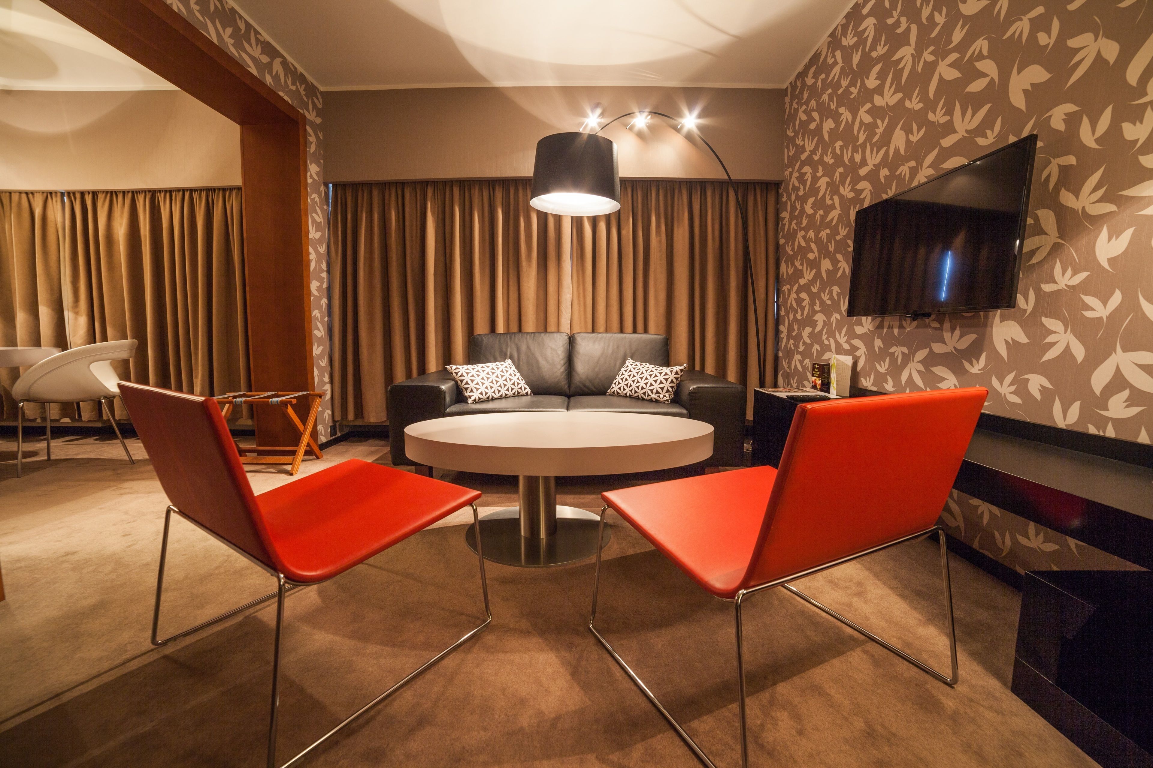 A modern hotel room with a round table, two red chairs, a sofa with patterned pillows, a flat-screen TV mounted on the wall, and a large window with curtains.