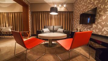 A modern hotel room with a round table, two red chairs, a sofa with patterned pillows, a flat-screen TV mounted on the wall, and a large window with curtains.