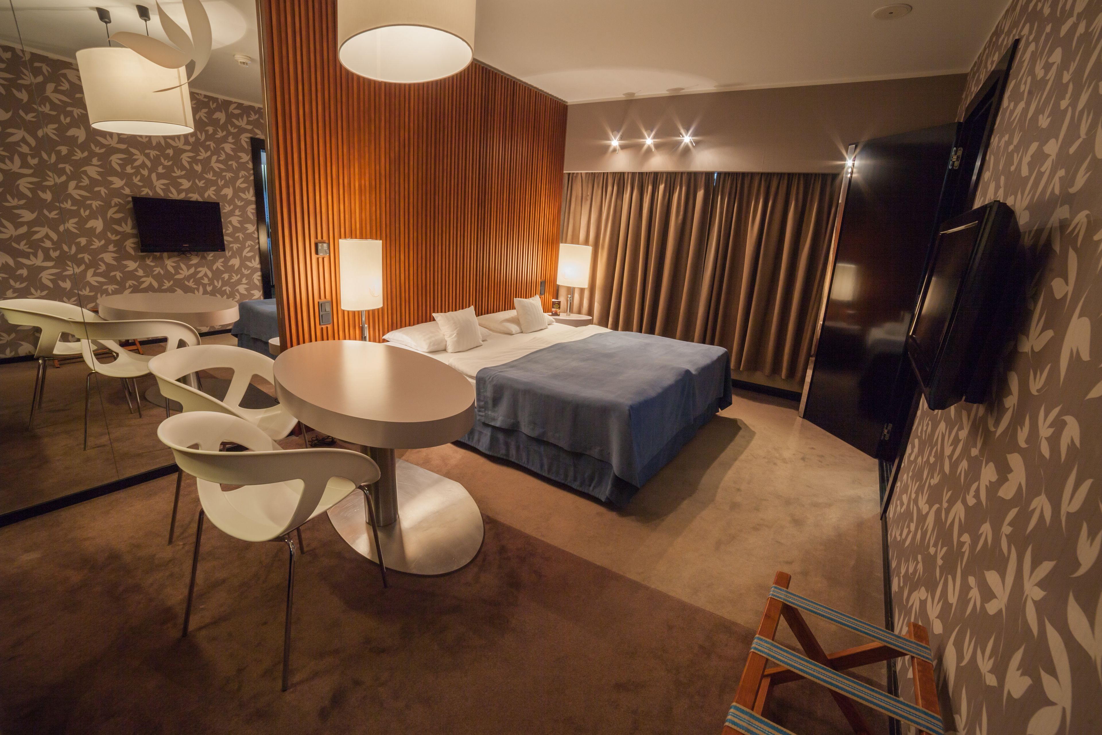 Una habitación de hotel moderna con una cama, dos mesitas de noche, un televisor de pantalla plana, una mesa de comedor con sillas y un gran ventanal con cortinas.