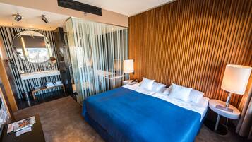 Quarto de hotel moderno com uma cama grande, banheiro separado com espelho e uma mesinha de cabeceira com luminária.