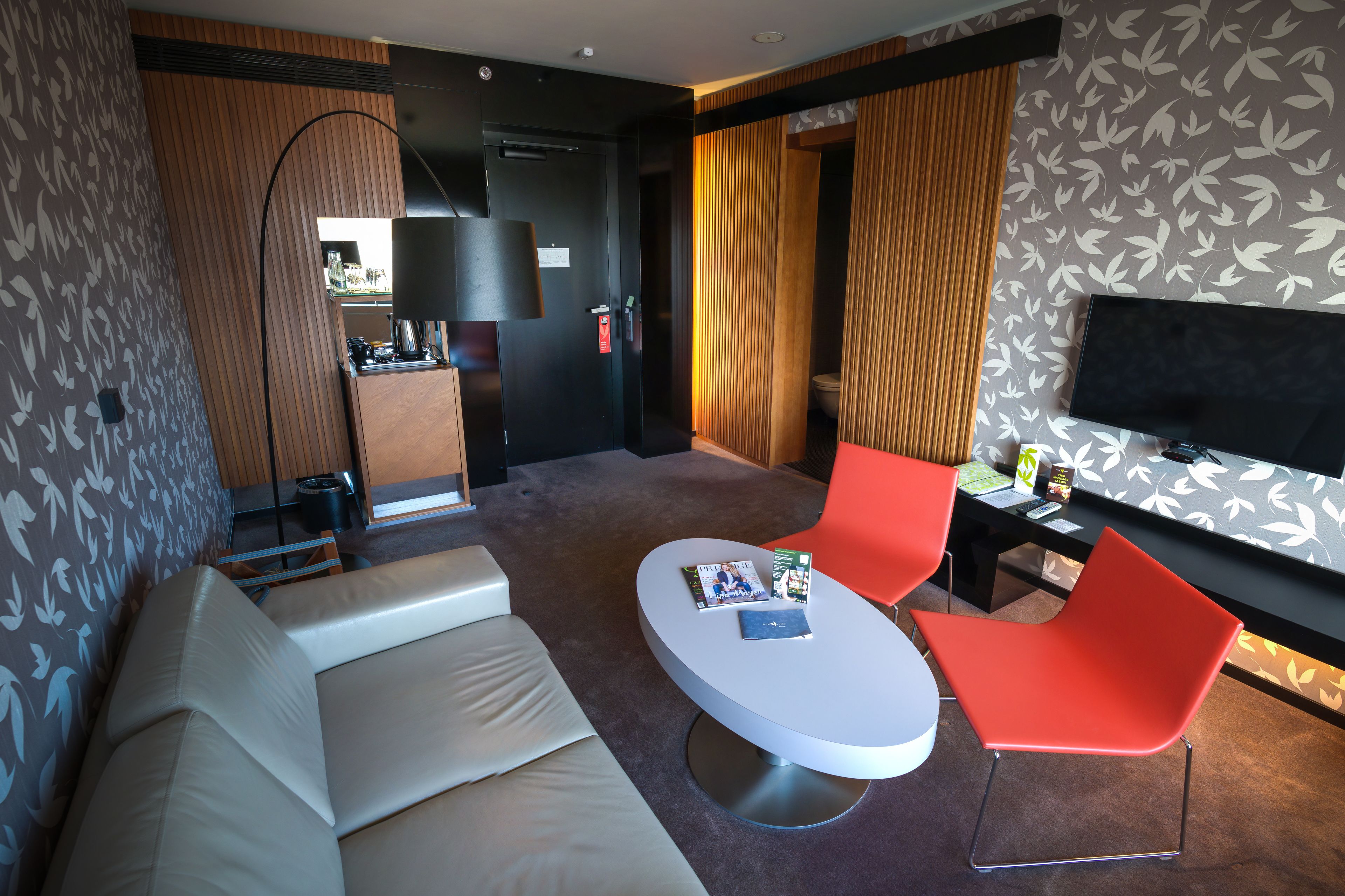 A modern hotel room with a sofa, a round table with magazines, a TV, and a desk with a chair.