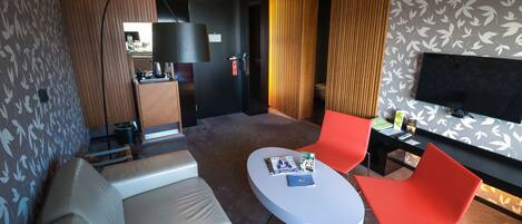 A modern hotel room with a sofa, a round table with magazines, a TV, and a desk with a chair.