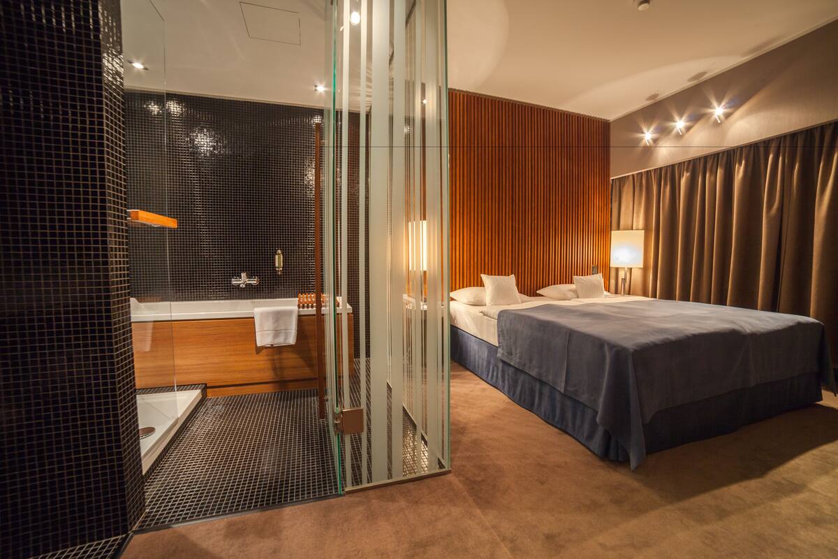 A modern hotel room with a glass-enclosed shower, a large bed with a blue bedspread, and a wooden panel wall.