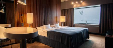 A hotel room with a bed, two bedside tables, a round table, and a view of the sky through the window.