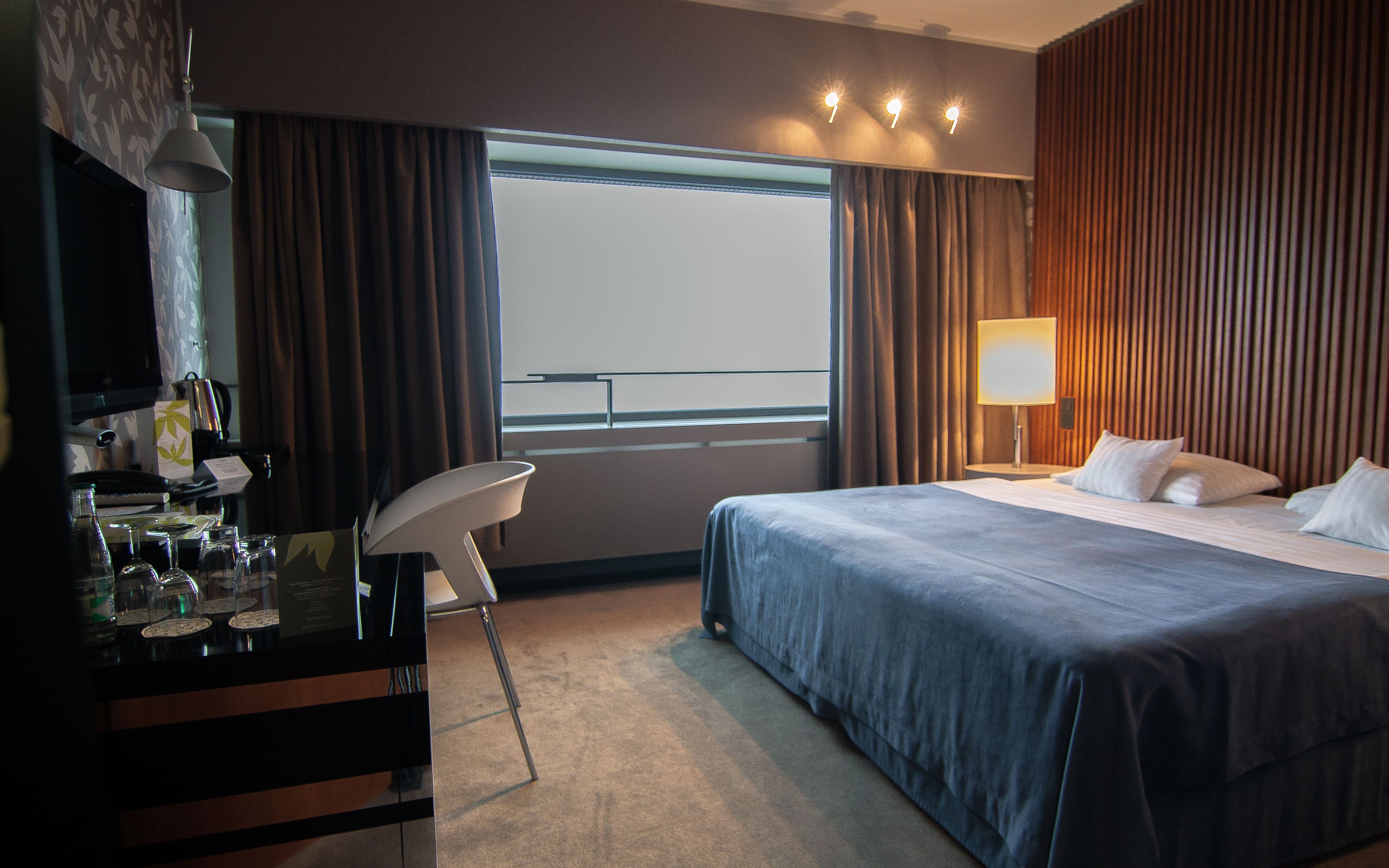 Una habitación de hotel con cama, escritorio con silla, lámpara y ventana con cortinas.