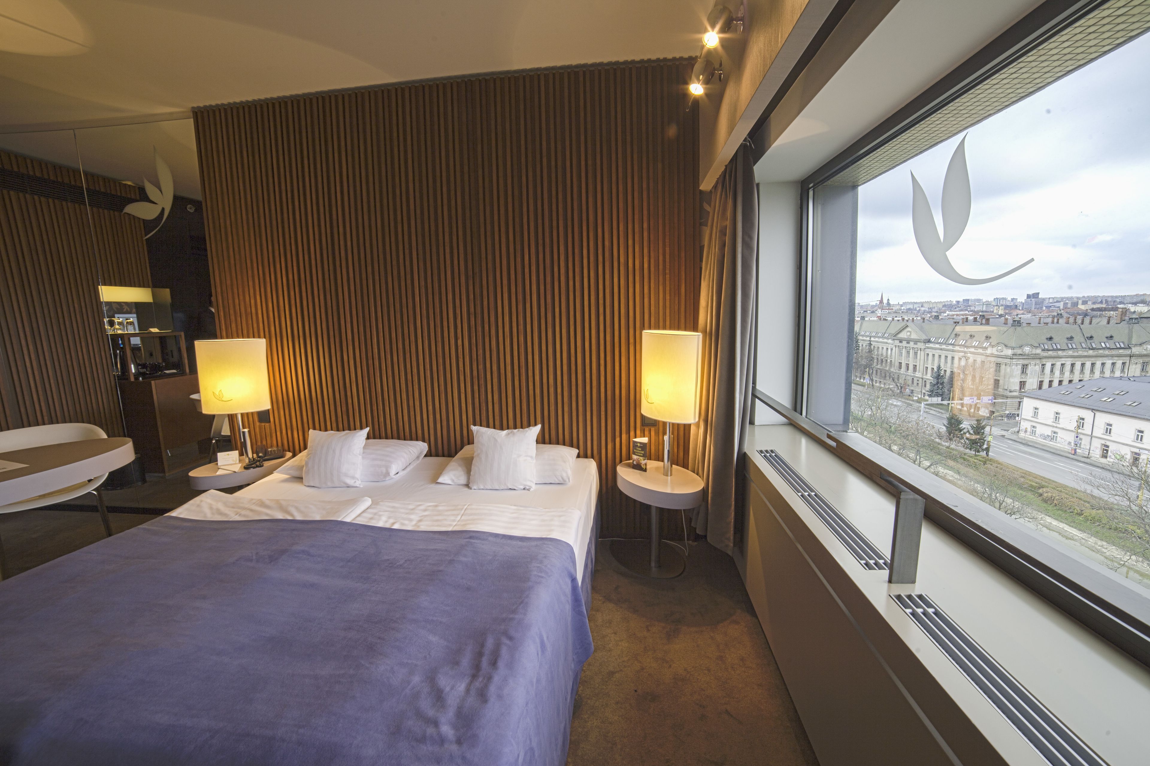 A hotel room with a large bed, two bedside lamps, a view of a cityscape through a window, and a mirror on the wall.