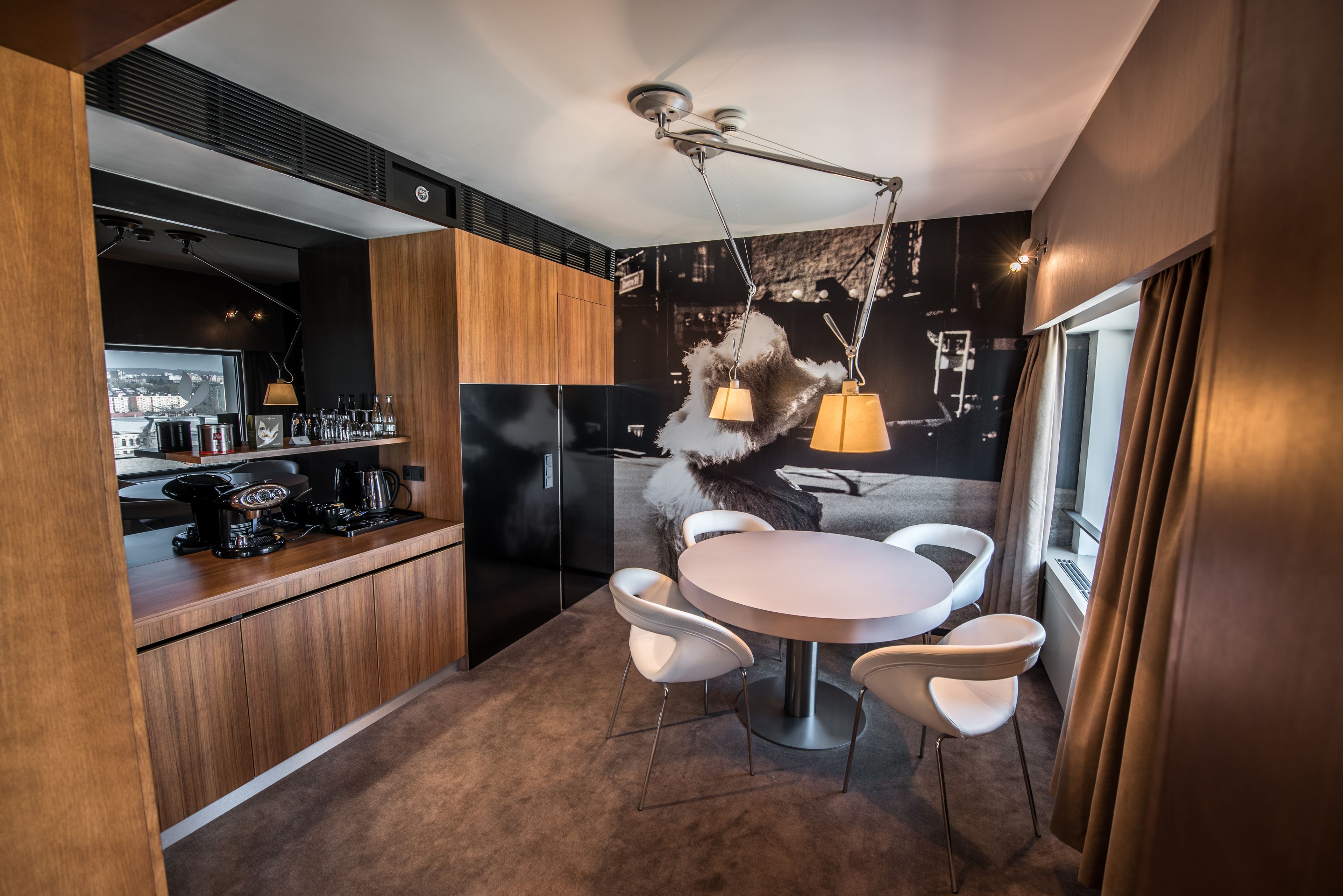 Cozinha moderna com mesa de jantar redonda e cadeiras brancas, área de bar com máquina de café e uma grande janela com cortinas.