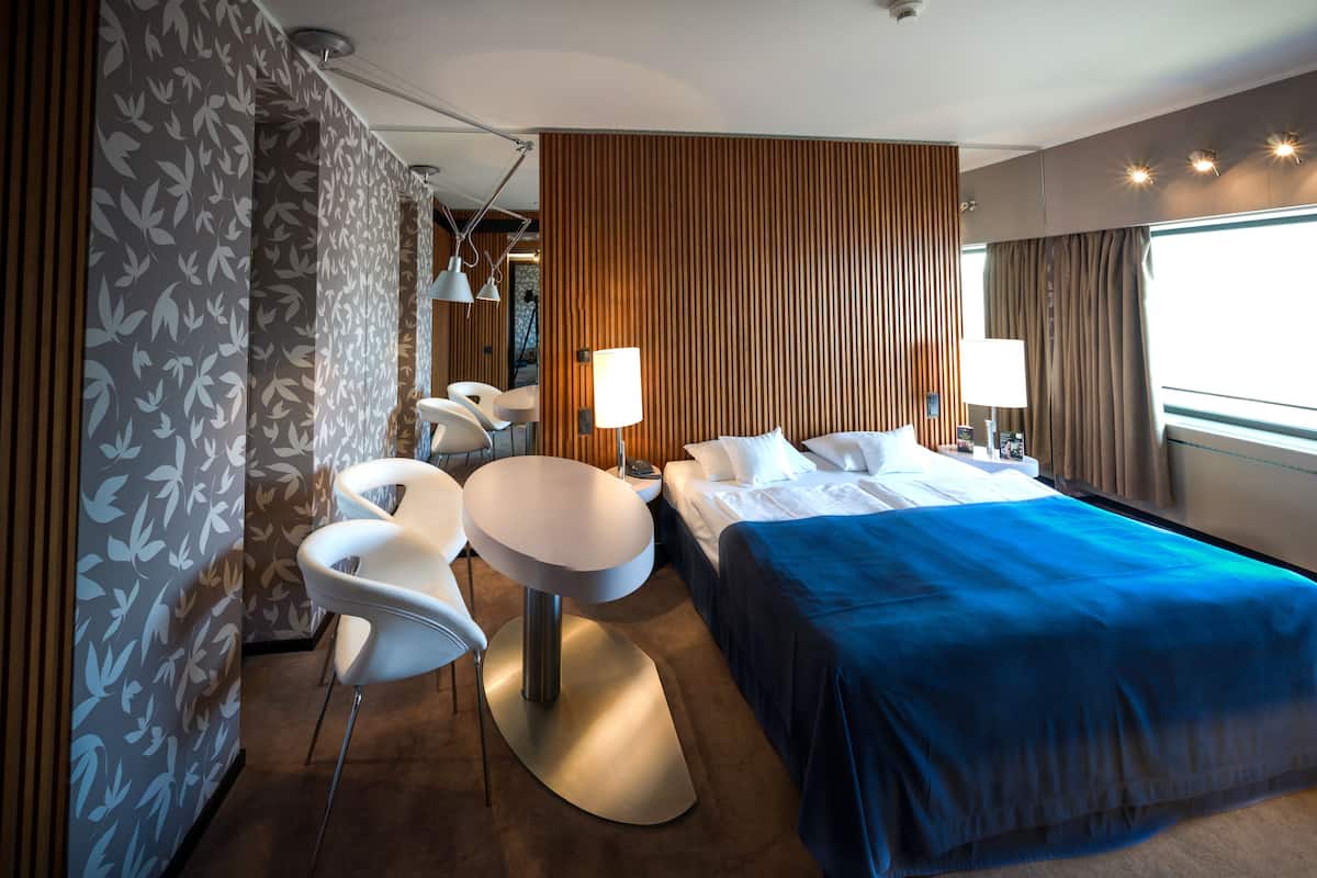 A modern hotel room with a large bed, two bedside tables, a round table with chairs, and a view of the city.