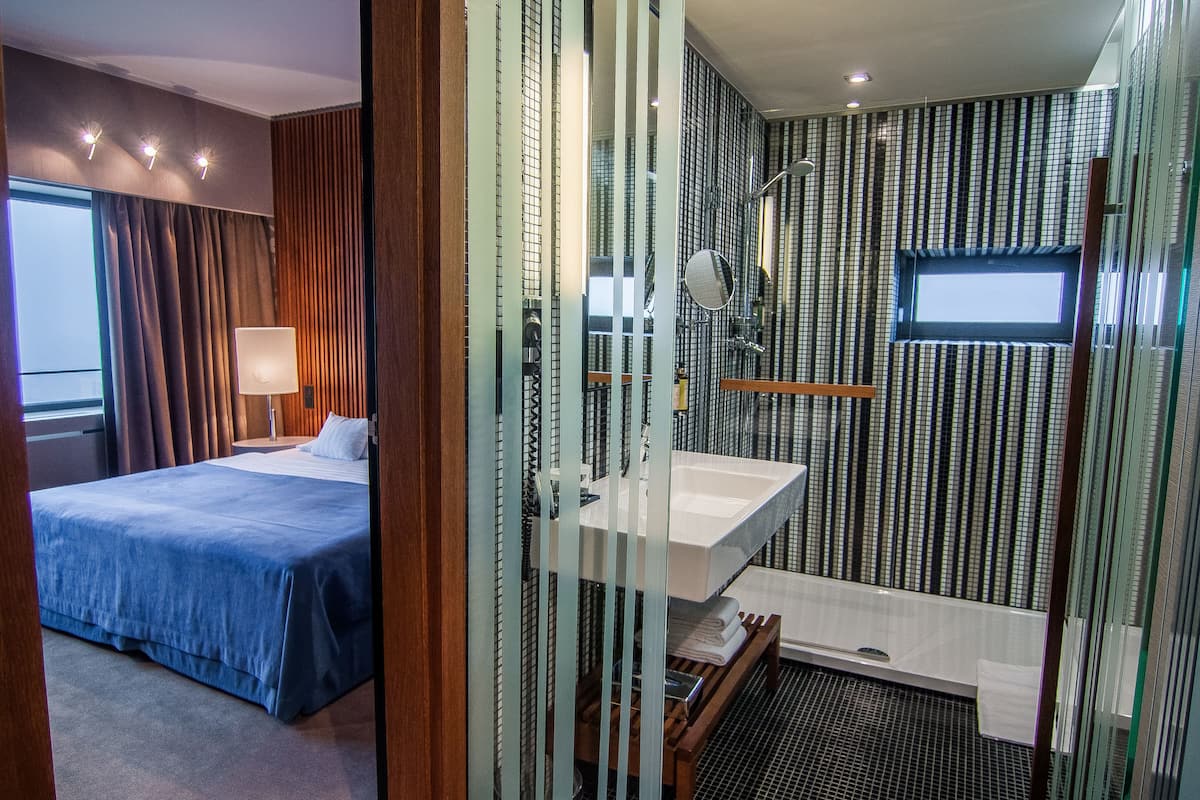 A hotel room with a bed, a bedside table with a lamp, a bathroom with a sink and mirror, and a shower area.