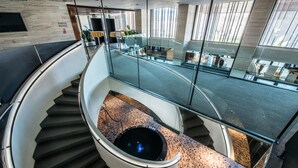 A modern interior with a curved glass staircase, a central circular feature, and a view of a well-lit area with furniture and plants. - Hotel Yasmin (Kosice)