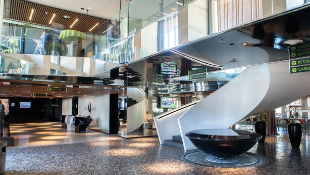 A modern hotel lobby with a glass atrium, a central spiral staircase, and directional signage for amenities. - Hotel Yasmin (Kosice)