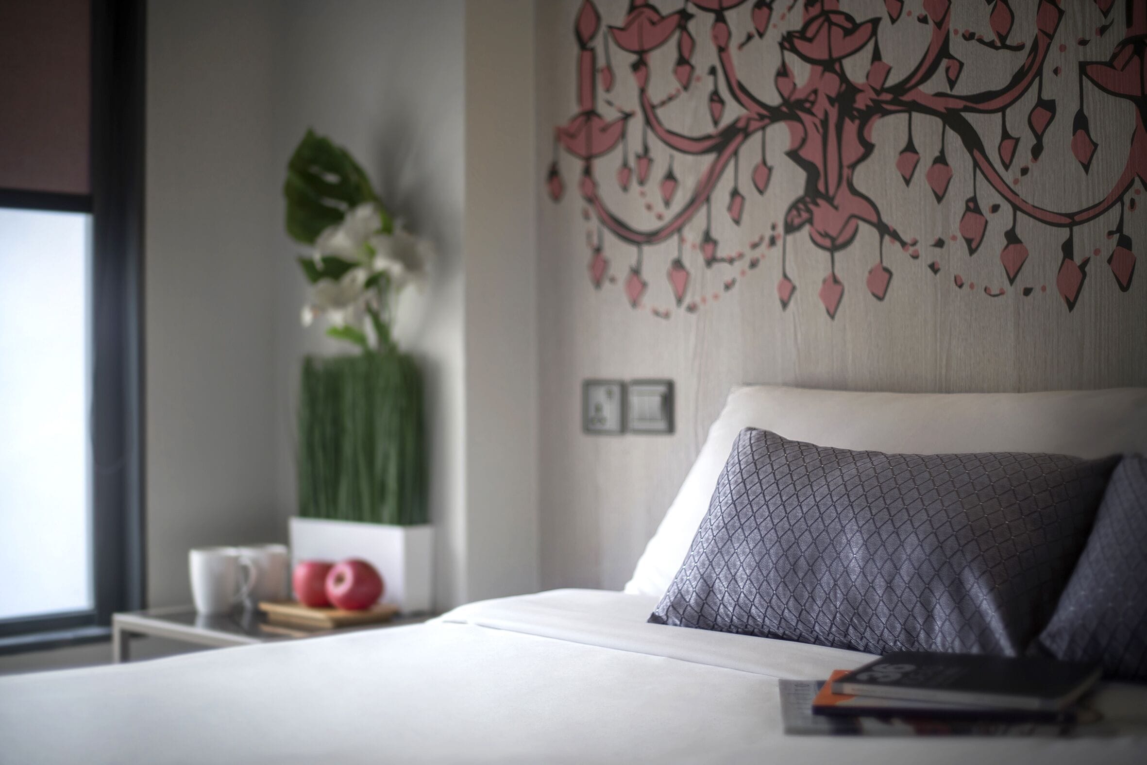 A neatly made bed with a white duvet and a grey patterned pillow, a bedside table with a book and a cup, and a wall with a decorative floral pattern.