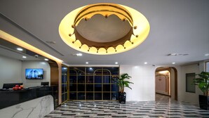 Un vestíbulo de hotel con una recepción, una lámpara de araña grande, un televisor mostrando el nombre del hotel y un mostrador de check-in.