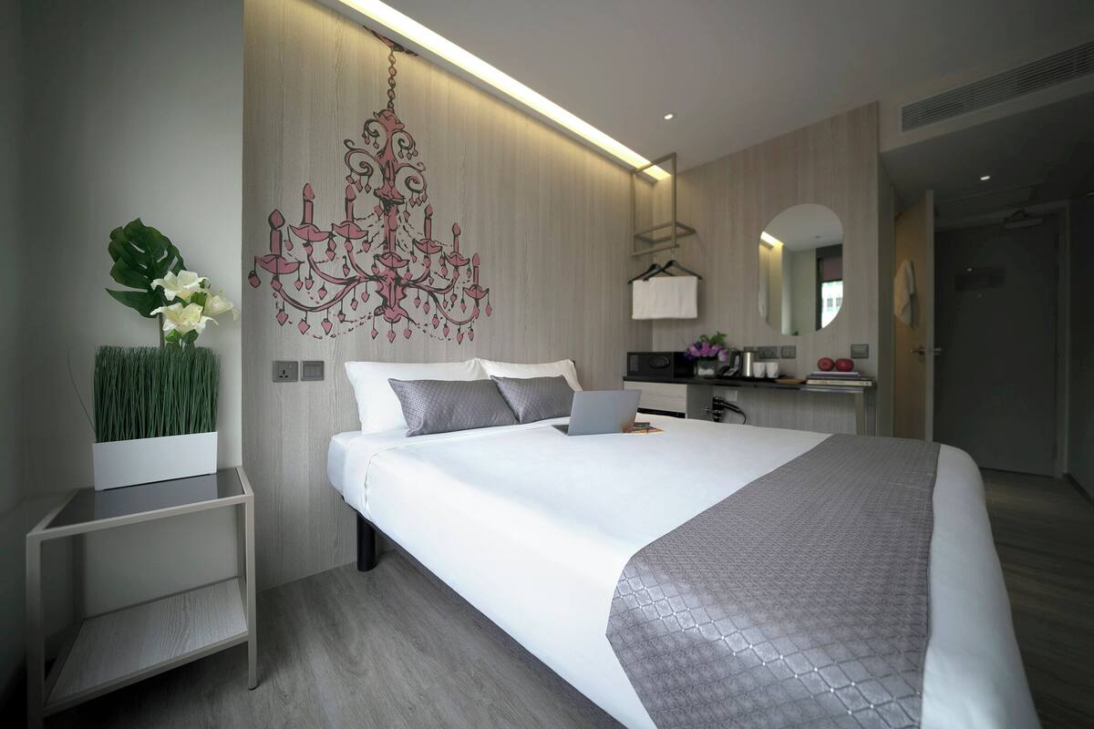 A modern hotel room with a large bed, bedside tables, a desk, a laptop, a chandelier, and a mirror.