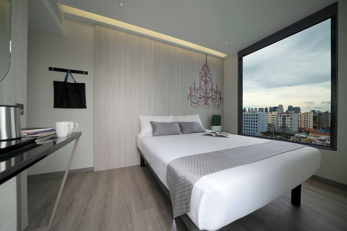 A modern hotel room with a large bed, a city view, a small desk with a cup and books, and a hanging bag.
