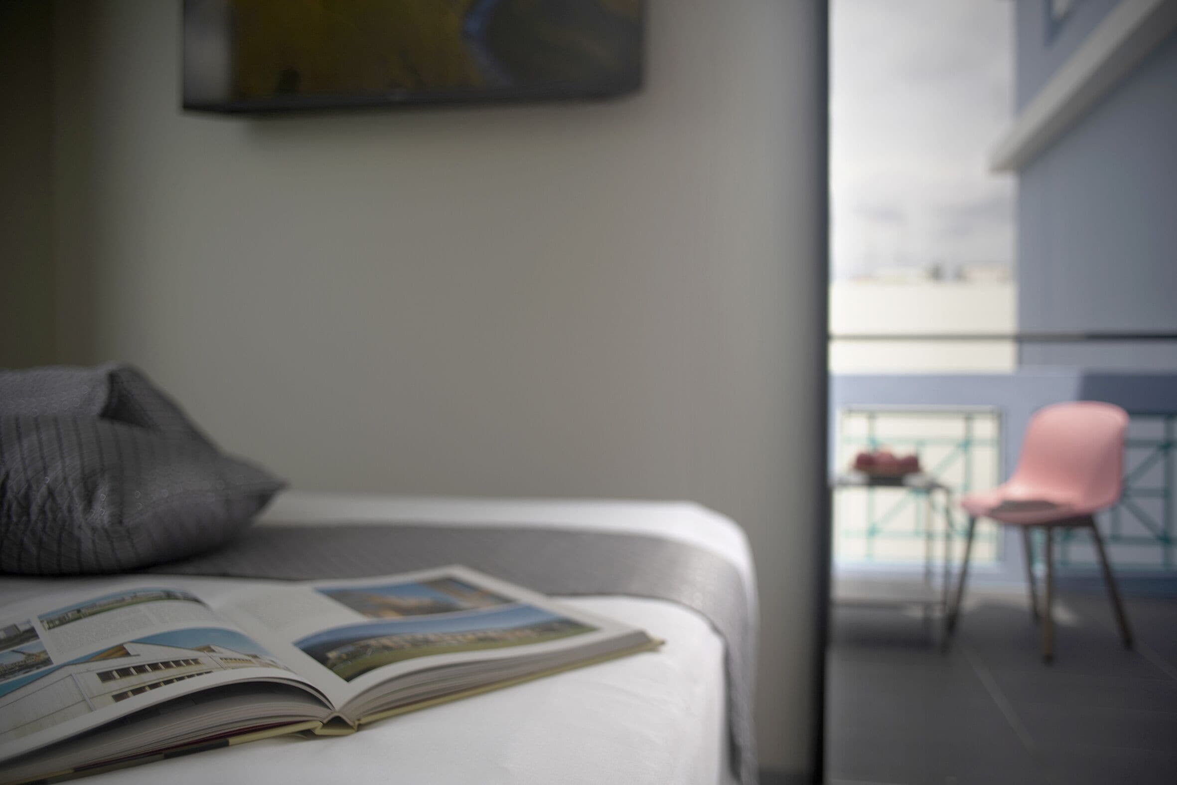 A hotel room with a bed, a magazine, a pillow, a pink chair, and a balcony with a view.