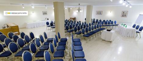 Un salón de conferencias con filas de sillas azules, una mesa blanca y un cartel para Grecian Castle Hotel.