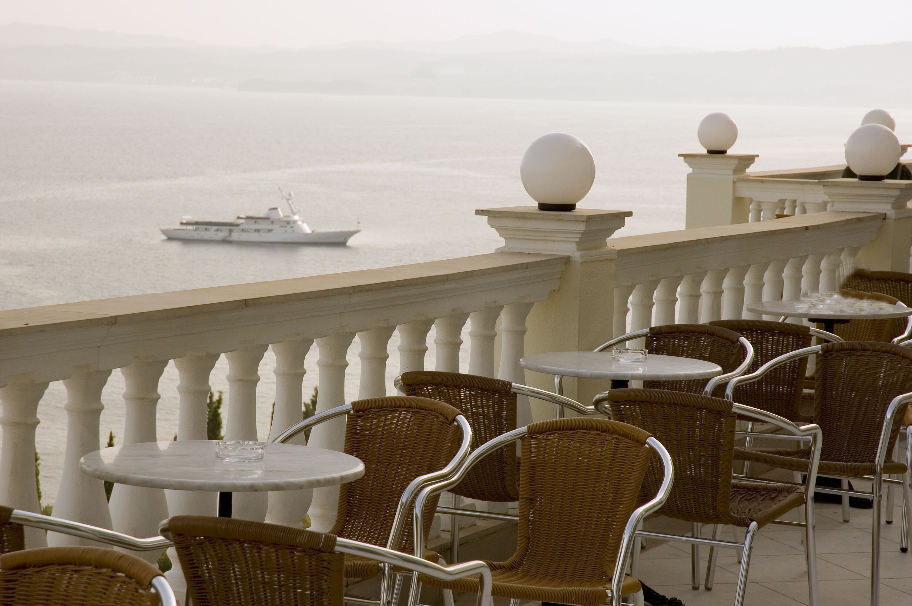 A seaside terrace with wicker chairs and tables, overlooking the sea and a ship.