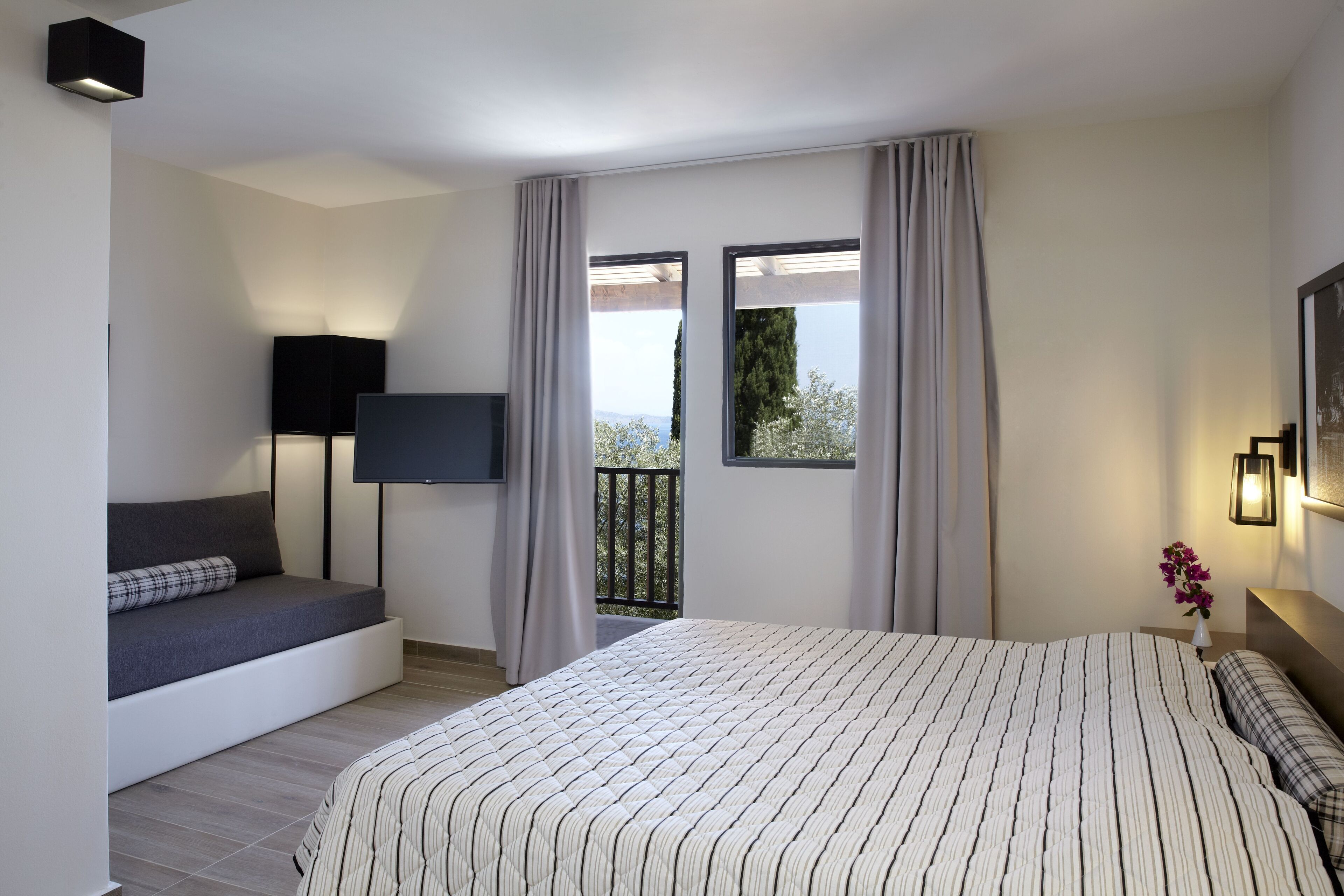 A modern bedroom with a large bed, a built-in bench, a flat-screen TV, and a balcony with curtains.