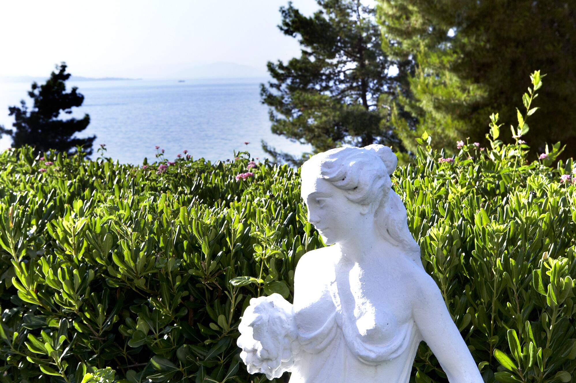 A white statue of a woman in a garden with green foliage and a view of the sea in the background.