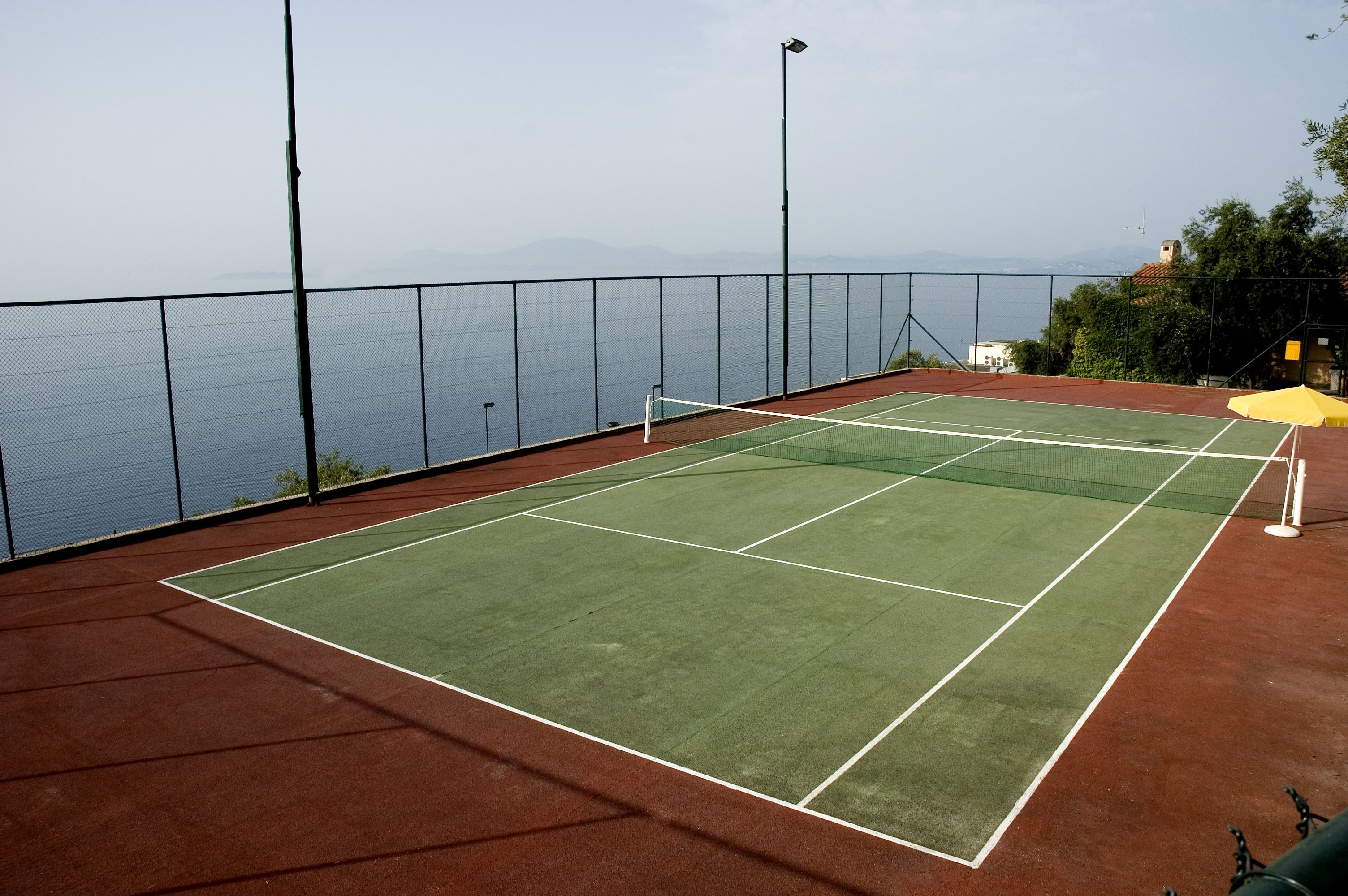 A tennis court with a net, surrounded by a fence, overlooking a body of water and hills.