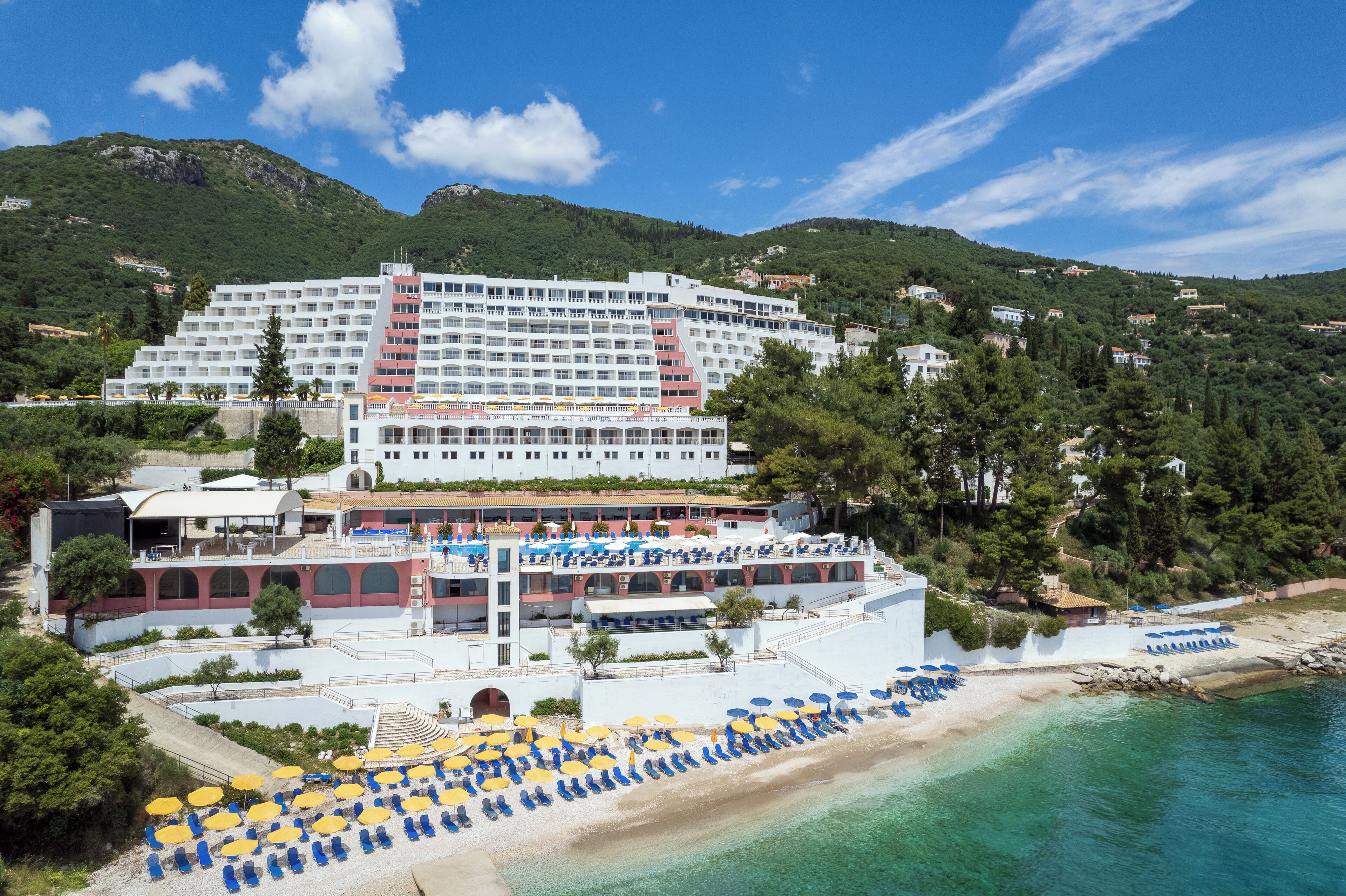 A large hotel with multiple floors, a beach with sun loungers and umbrellas, and a mountainous backdrop.