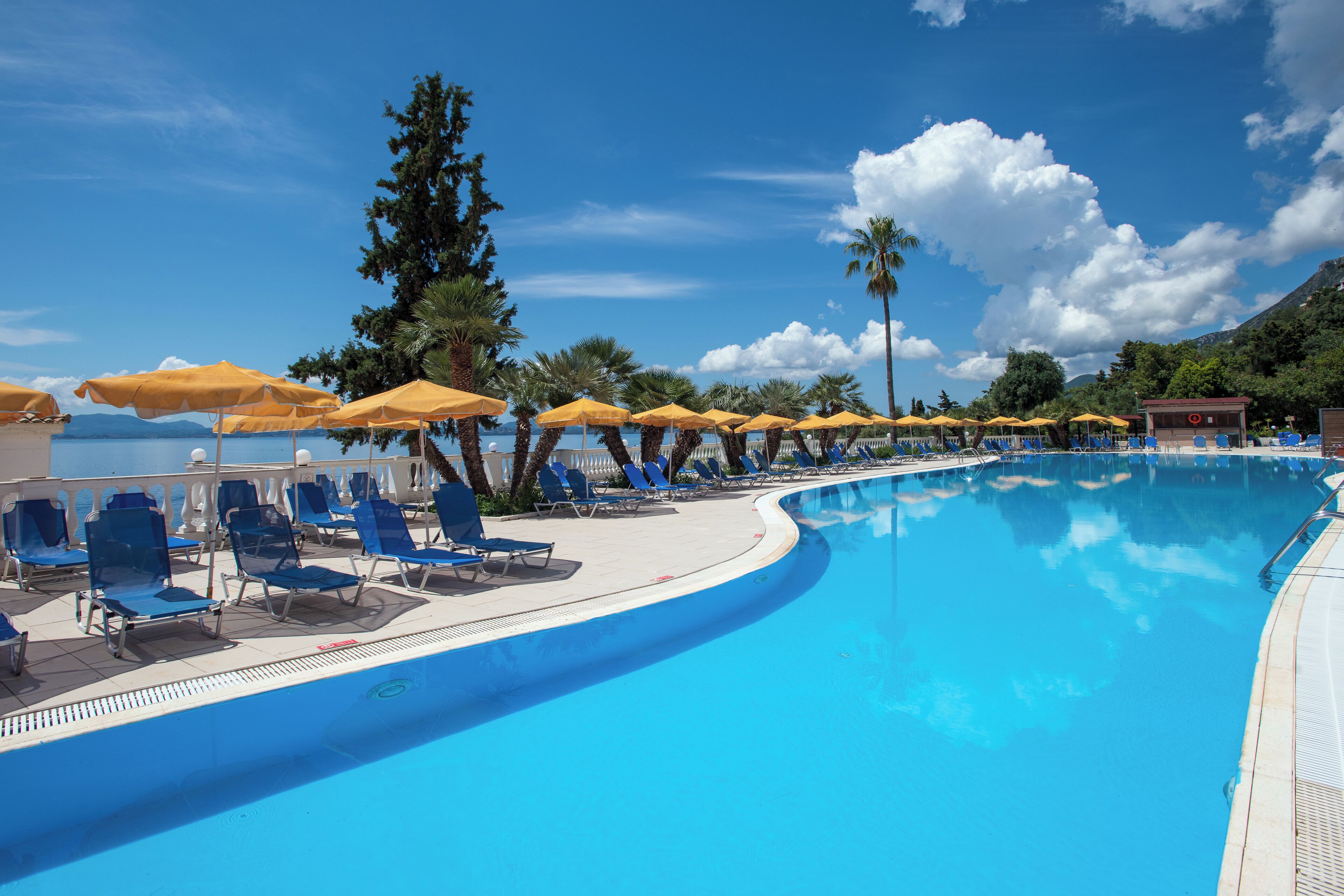 A poolside area with blue lounge chairs and yellow umbrellas, surrounded by palm trees and a clear blue sky.