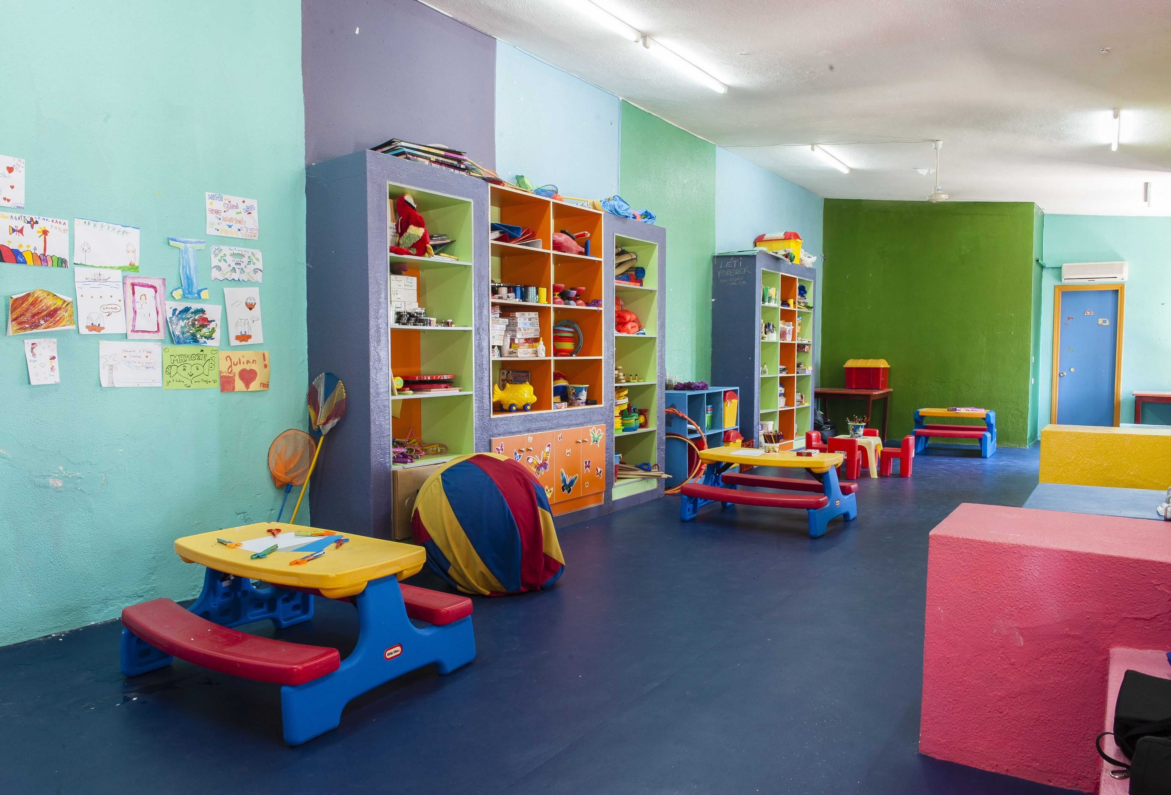 Een kleurrijke kinderspeelkamer met tafels, stoelen en planken vol speelgoed en boeken.