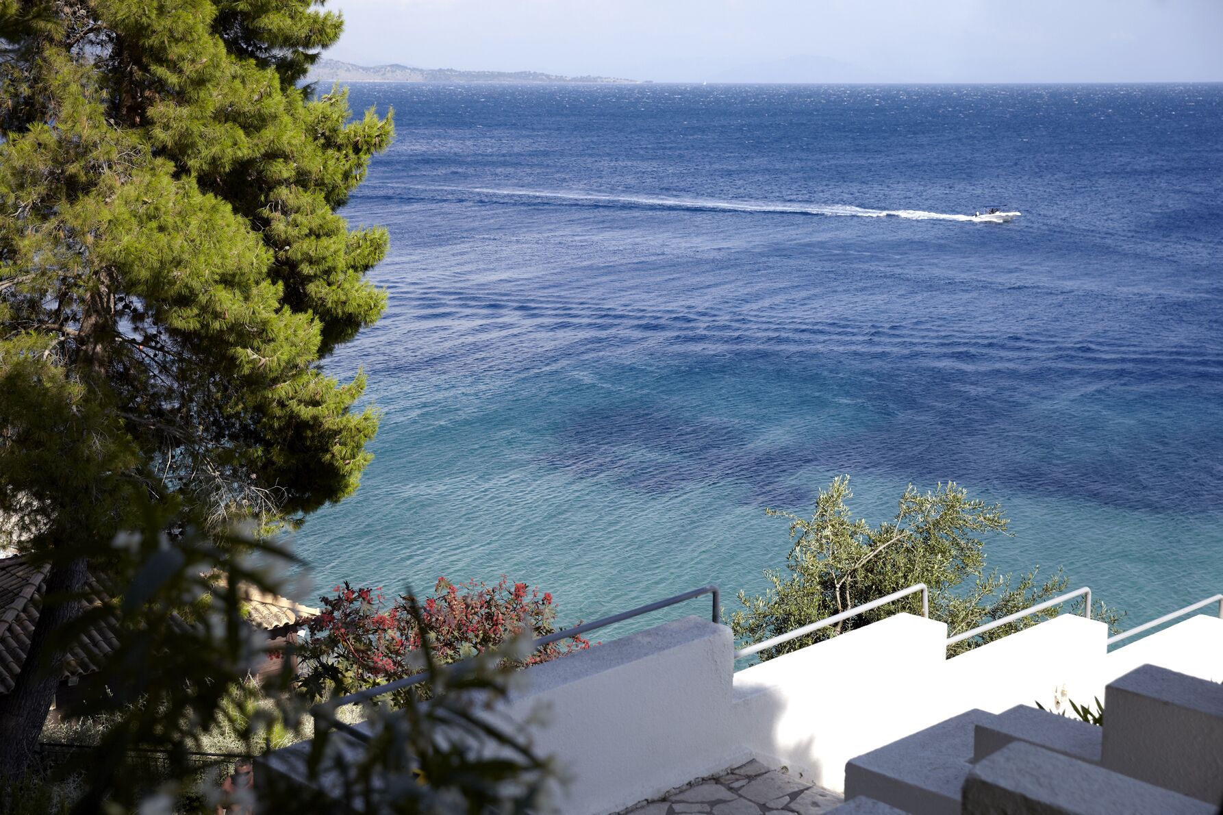 Vista costera con un mar azul y transparente, un barco a lo lejos y una escalera que baja hacia el agua.
