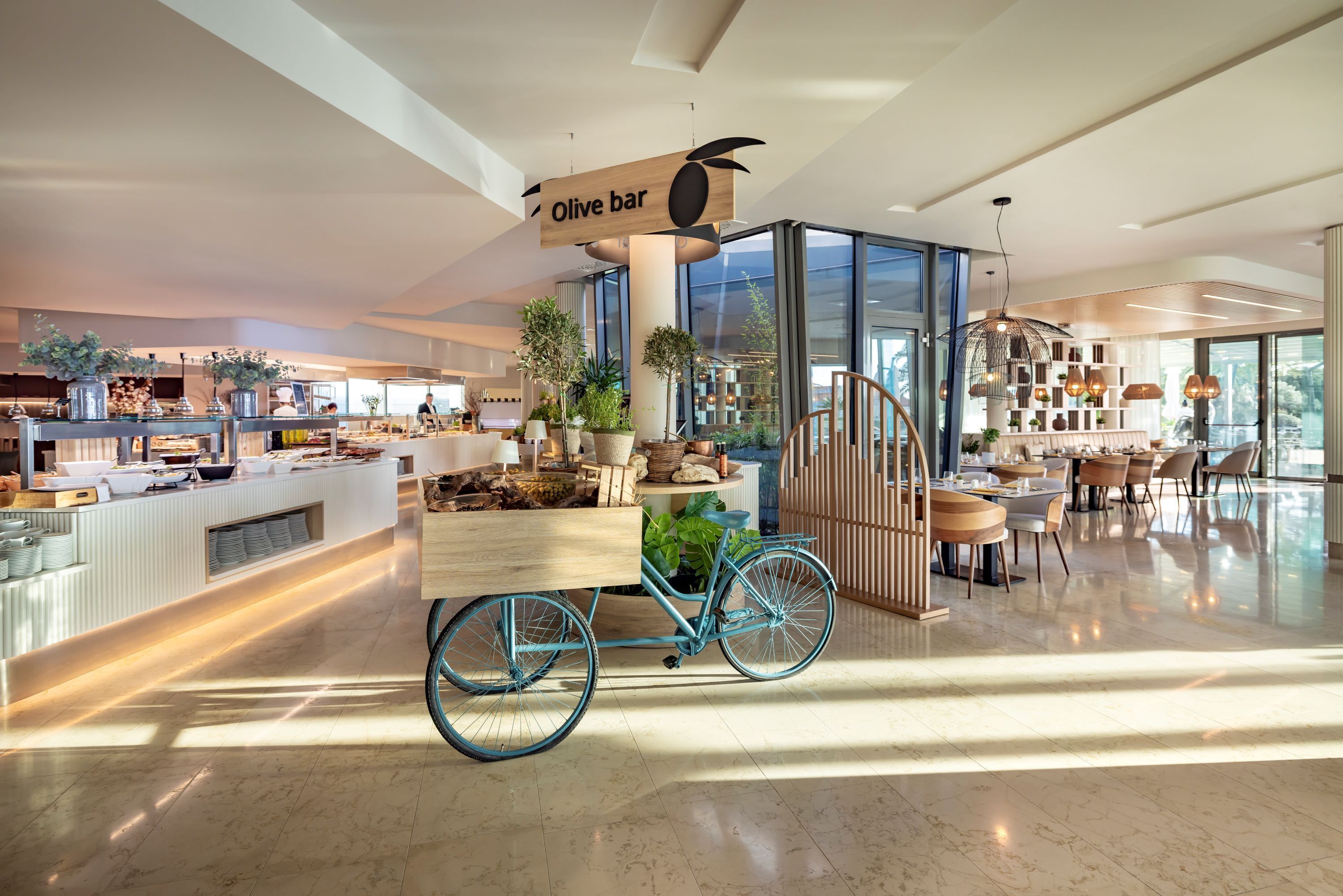 Un moderno vestíbulo de hotel con una barra central identificada como 'Olive bar', que incluye un carrito de bicicleta, plantas en macetas y una escalera que conduce a otro nivel.