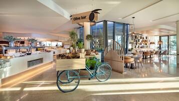 A modern hotel lobby with a central bar area labeled 'Olive bar', featuring a bicycle cart, potted plants, and a staircase leading to another level.