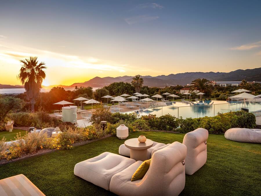 Área da piscina com espreguiçadeiras, guarda-sóis e vista para o pôr do sol sobre as montanhas.